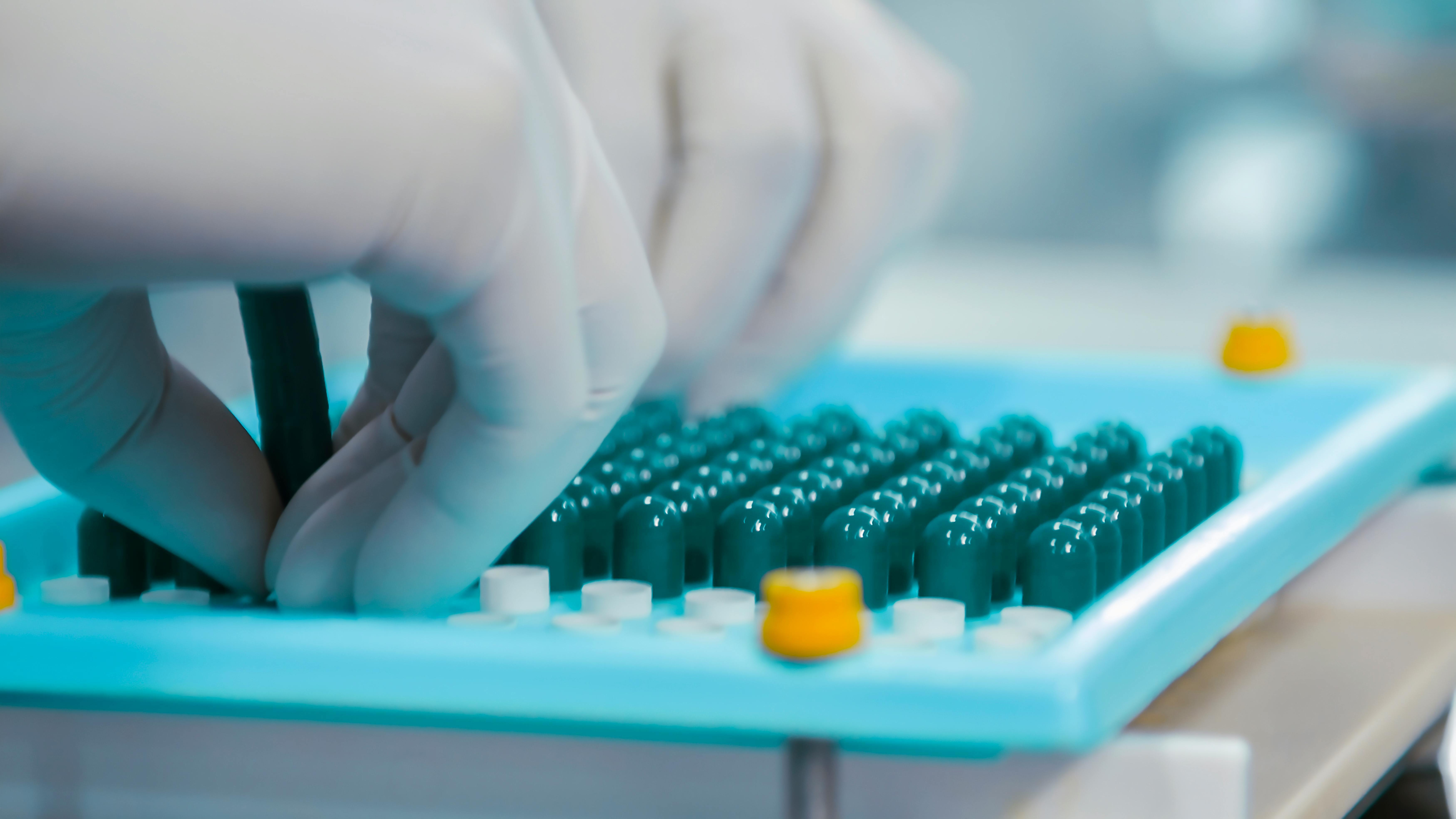 Close-up of gloved hands arranging small capsules or components in a sterile laboratory tray, manufacturing of pharmaceutical production.